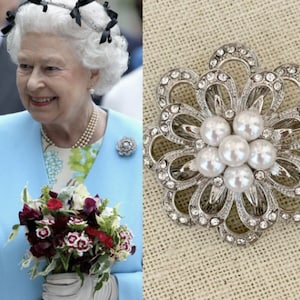 May include: A silver-toned flower-shaped brooch with faux pearls and clear crystals. The brooch has a pin backing and is set against a neutral background. The brooch is a jewelry item.