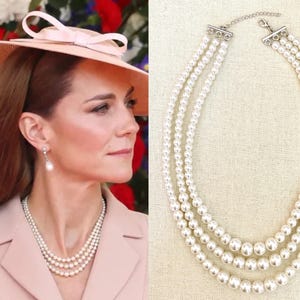 May include: A three-strand pearl necklace with a silver clasp. The necklace features graduated sizes of round, white pearls. The necklace is shown on a cream-coloured background. The necklace is a classic piece of jewellery.