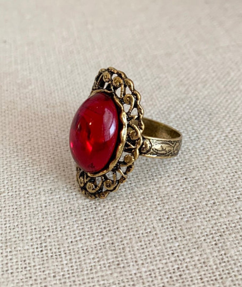 May include: A gold ring with an ornate design and a large, oval-shaped red gemstone.