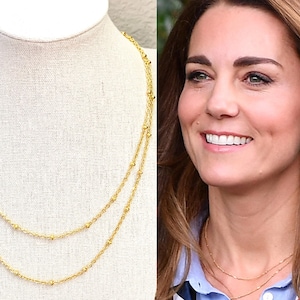May include: A double-layered gold necklace with delicate beaded accents. The necklace is displayed on a white mannequin head. The image also shows a woman wearing a similar necklace, with a blue shirt and hoop earrings.