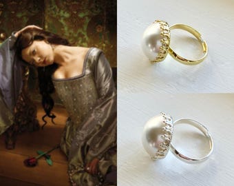 May include: Two rings with large, white, pearl-like stones. One ring has a gold band and decorative gold setting, the other has a silver band and setting. The rings are on a white surface. A painting of a woman in a gown is in the background.