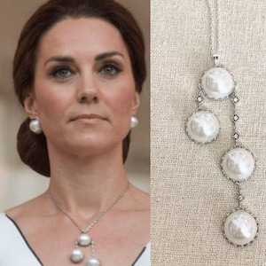 May include: A silver necklace with a long pendant featuring four white pearl-like orbs. The pendant is adorned with small silver accents. The necklace is displayed on a white fabric background.