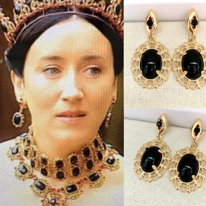 May include: A pair of gold earrings with black oval stones set in a filigree design. The earrings are dangling and have a red stone at the top.