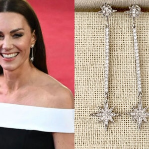 May include: A pair of silver-toned, long dangle earrings. Each earring features a starburst design at the top and bottom, connected by a straight line of small, sparkling stones. The earrings are displayed against a neutral background.