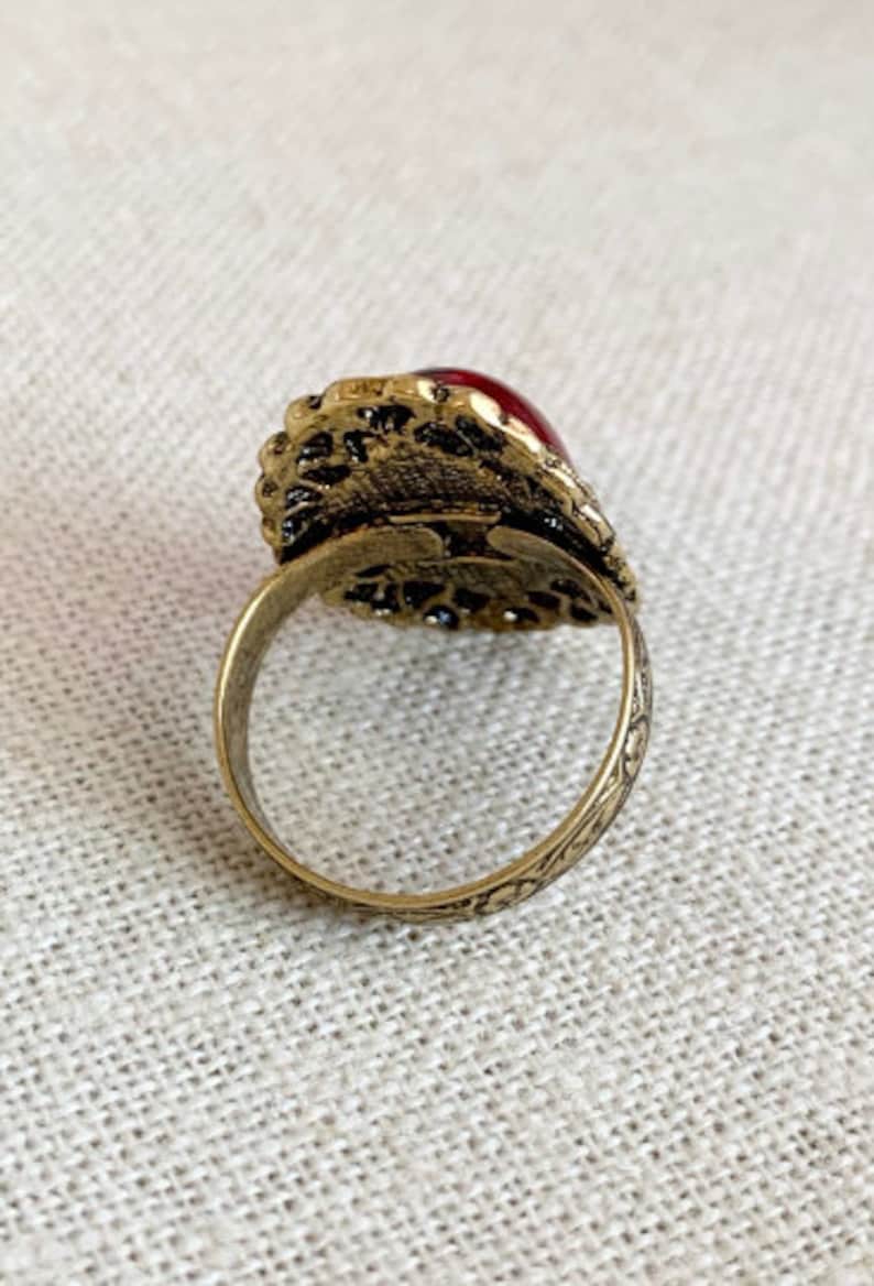 May include: A gold ring with an ornate design and a large red gemstone. The ring is set on a white fabric background.