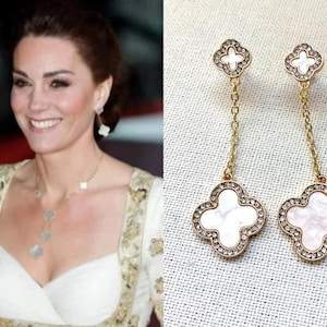 May include: A pair of gold-tone drop earrings featuring a small clover-shaped stud connected by a delicate chain to a larger clover-shaped pendant. Each clover element is inlaid with white mother-of-pearl and bordered by small, clear, sparkling crystals.
