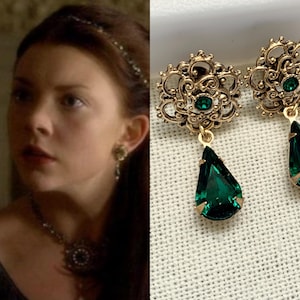 May include: A pair of ornate gold-tone earrings with emerald green teardrop-shaped pendants. The earrings feature a filigree design with a small green gem in the center of the floral-shaped top. The pendants are a deep, rich green.