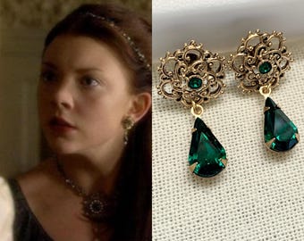May include: A pair of ornate gold-tone earrings with emerald green teardrop-shaped pendants. The earrings feature a filigree design with a small green gem in the center of the floral-shaped top. The pendants are a deep, rich green.
