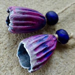 Puede incluir: Dos cuentas hechas a mano en forma de campana con un diseño texturizado y acanalado. Las cuentas están pintadas con tonos de morado, rosa y azul, con un acento blanco en los bordes. Cada cuenta está conectada a una pequeña cuenta azul oscuro y una cuerda de fibra natural.