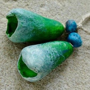 May include: Two green, hollow, teardrop-shaped beads with a light blue-green wash, connected to two smaller blue beads and a natural fiber string. The beads rest on a light tan, textured surface.