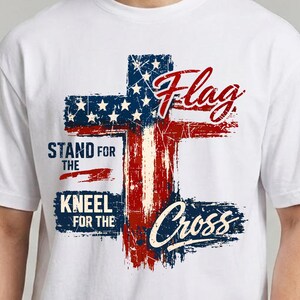 May include: White t-shirt featuring a distressed American flag cross design. The design incorporates red and blue elements with the words "Flag", "Stand for the", "Kneel for the", and "Cross".