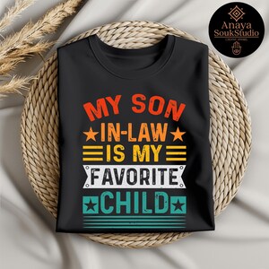 May include: Black t-shirt with the text "MY SON IN-LAW IS MY FAVORITE CHILD" in orange, yellow, and white. The design includes stars and horizontal lines. The shirt is folded on a woven surface.