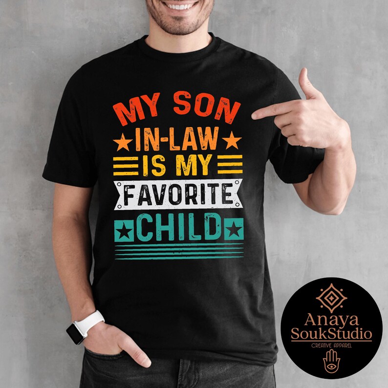 May include: Black t-shirt with the text "MY SON IN-LAW IS MY FAVORITE CHILD" in orange, yellow, white, and teal. The shirt has a crew neck and short sleeves. The design includes stars and horizontal lines.
