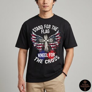 K&ouml;nnte beinhalten: Schwarzes T-Shirt mit einem grafischen Design. Das Design beinhaltet ein Kreuz, Fl&uuml;gel und Elemente der amerikanischen Flagge in Rot, Wei&szlig; und Blau. Der Text lautet "STAND FOR THE FLAG" und "KNEEL FOR THE CROSS".