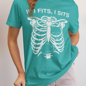 May include: A teal short-sleeved t-shirt featuring a white graphic print of a human rib cage with a small cat sitting inside the chest cavity. Above the rib cage, the text reads "IF I FITS, I SITS" in a bold, white, sans-serif font.