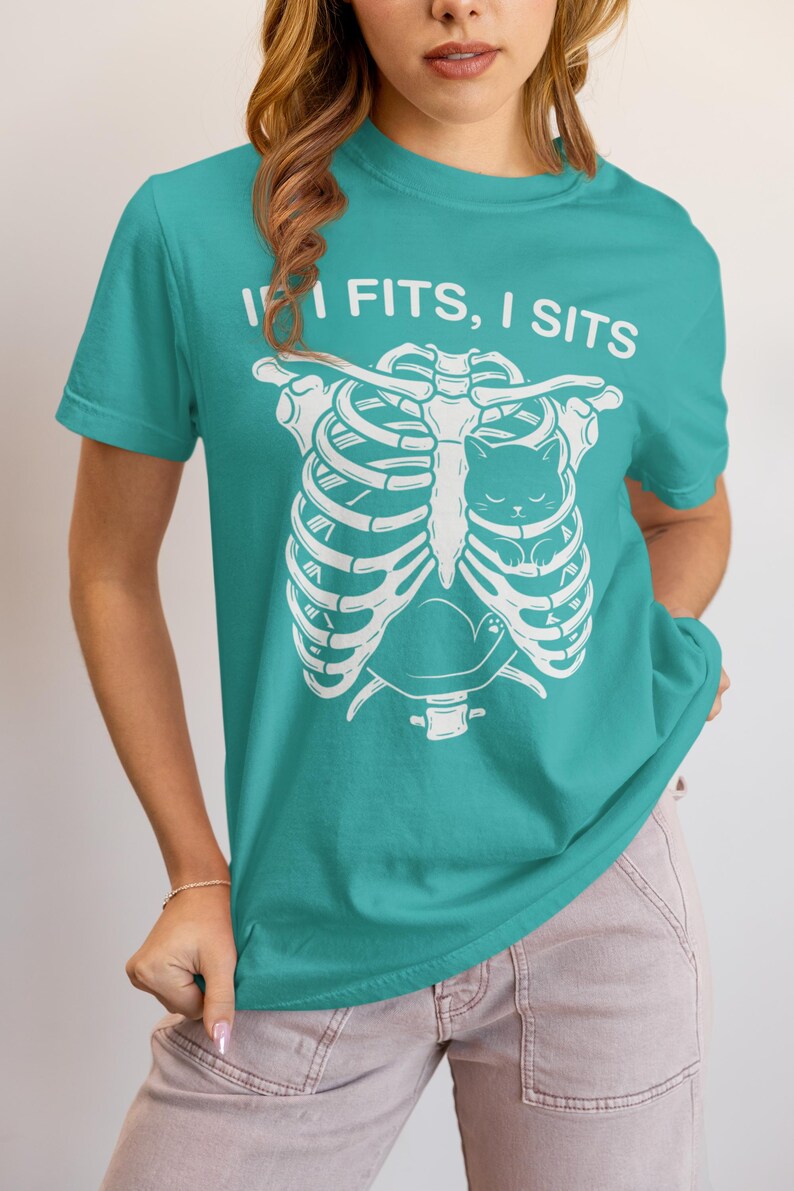 May include: A teal short-sleeved t-shirt featuring a white graphic print of a human rib cage with a small cat sitting inside the chest cavity. Above the rib cage, the text reads "IF I FITS, I SITS" in a bold, white, sans-serif font.