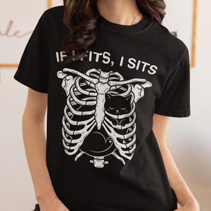 May include: A black short-sleeved t-shirt featuring a white graphic of a human rib cage with a small black cat curled up inside. Above the graphic, the text reads IF I FITS, I SITS in a white sans-serif font. The shirt has a crew neckline.