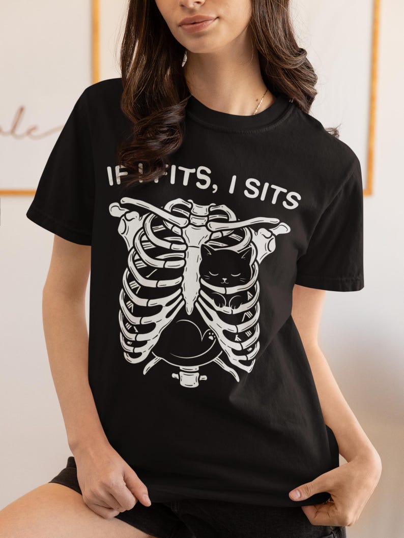 May include: A black short-sleeved t-shirt featuring a white graphic of a human rib cage with a small black cat curled up inside. Above the graphic, the text reads IF I FITS, I SITS in a white sans-serif font. The shirt has a crew neckline.