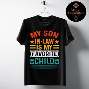 May include: Black t-shirt with the text "MY SON IN-LAW IS MY FAVORITE CHILD" in orange, yellow, and teal. The design includes stars and horizontal lines. The shirt is hanging on a metal hanger.
