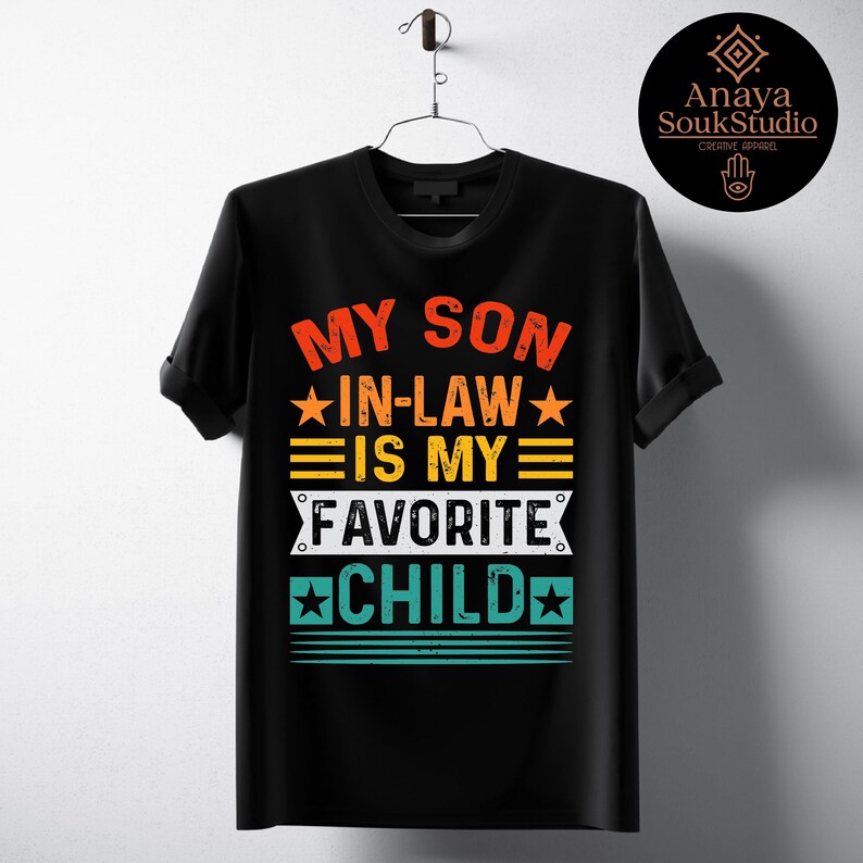May include: Black t-shirt with the text "MY SON IN-LAW IS MY FAVORITE CHILD" in orange, yellow, and teal. The design includes stars and horizontal lines. The shirt is hanging on a metal hanger.