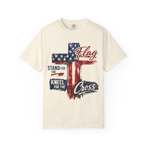 Puede incluir: Camiseta de color crema con un dise&ntilde;o de bandera estadounidense desgastada en rojo, blanco y azul. El dise&ntilde;o incluye las palabras "Flag", "Stand for the", "Kneel for the" y "Cross".