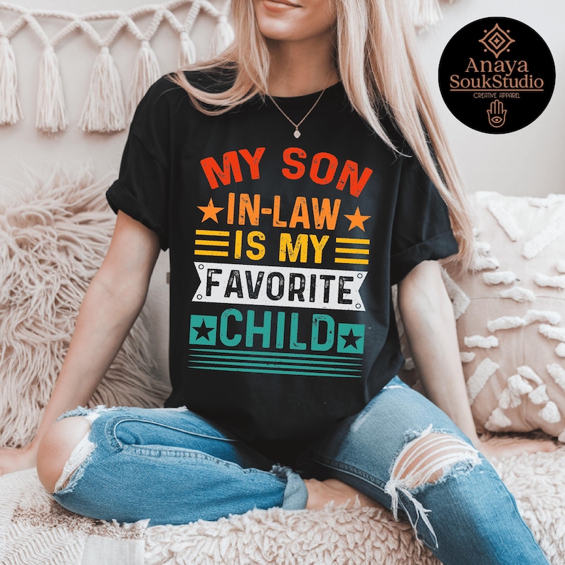 May include: Black t-shirt with the text "MY SON IN-LAW IS MY FAVORITE CHILD" in a retro design. The text is in red, yellow, orange, white, and green. The shirt is worn by a person wearing blue jeans.