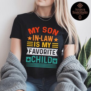 May include: Black t-shirt with the text "MY SON IN-LAW IS MY FAVORITE CHILD" in orange, yellow, and white. The design includes stars and horizontal stripes. A round logo is visible in the upper right corner.