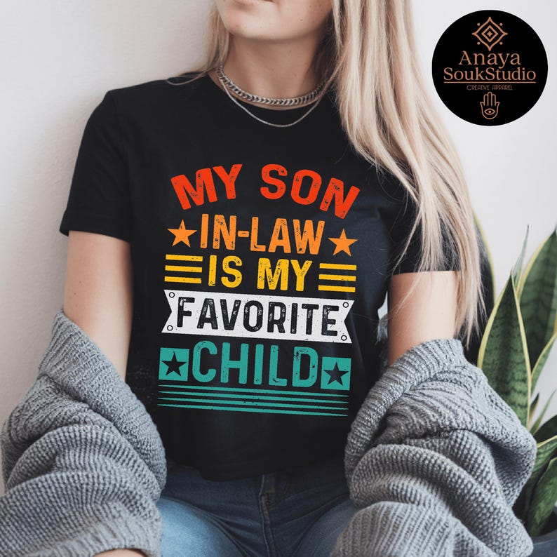 May include: Black t-shirt with the text "MY SON IN-LAW IS MY FAVORITE CHILD" in orange, yellow, and white. The design includes stars and horizontal stripes. A round logo is visible in the upper right corner.