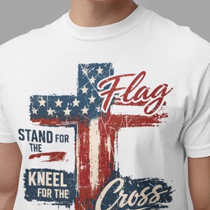 Puede incluir: Camiseta blanca con un dise&ntilde;o de cruz desgastada en los colores de la bandera americana. El dise&ntilde;o incluye la palabra "Flag" y las frases "Stand for the" y "Kneel for the Cross."