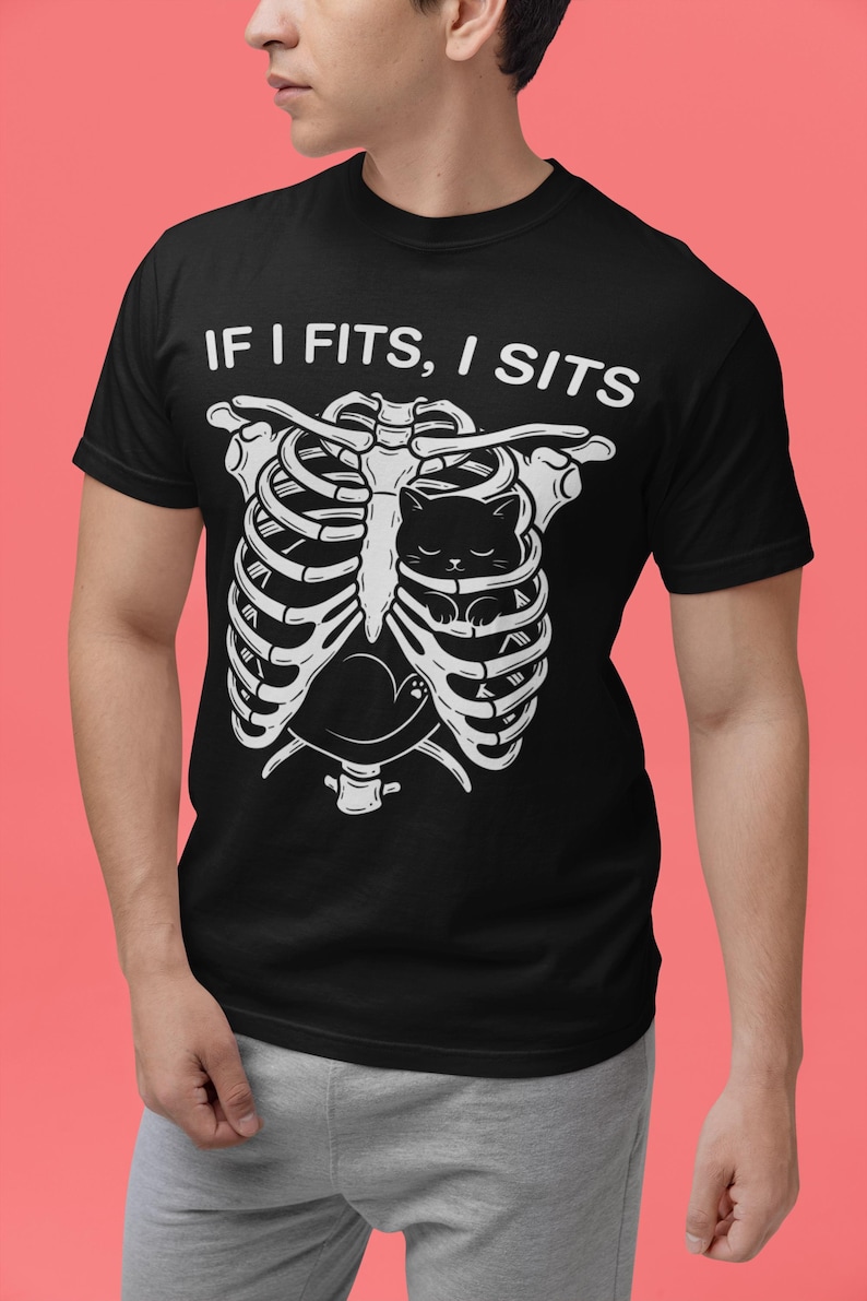 May include: A black short-sleeved t-shirt featuring a white graphic of a human rib cage with a small black cat curled up inside. Above the rib cage, the text reads IF I FITS, I SITS in white capital letters. The shirt has a crew neckline.