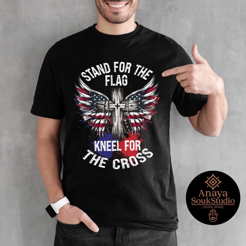 K&ouml;nnte beinhalten: Schwarzes T-Shirt mit einem patriotischen Design mit einem Kreuz, Fl&uuml;geln der amerikanischen Flagge und dem Text "STAND FOR THE FLAG, KNEEL FOR THE CROSS". Das Shirt hat auch ein kleines Logo von Anaya SoukStudio.