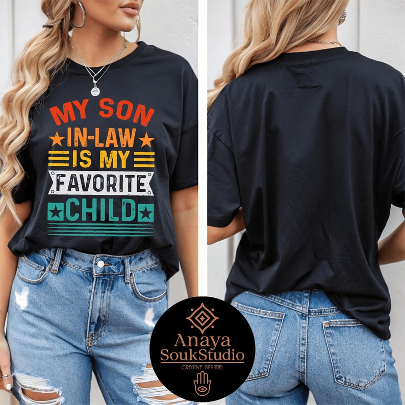 May include: Black t-shirt with the text "MY SON IN-LAW IS MY FAVORITE CHILD" in orange, yellow, green, and white. The shirt has a crew neck and short sleeves. The back of the shirt is plain black. The shirt is paired with blue jeans.