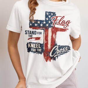 Puede incluir: Camiseta blanca con un dise&ntilde;o de cruz de la bandera estadounidense desgastada. La camiseta incluye las palabras "Flag" y "Cross" en escritura cursiva, junto con "Stand for the" y "Kneel for the".