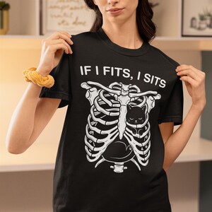 May include: A black short-sleeved t-shirt featuring a white graphic of a human rib cage with a small black cat nestled inside. Above the graphic, the text reads IF I FITS, I SITS in a bold white font. The shirt has a crew neckline and a standard fit.