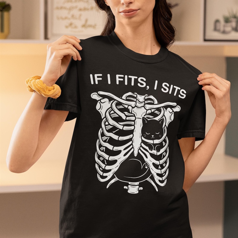 May include: A black short-sleeved t-shirt featuring a white graphic of a human rib cage with a small black cat nestled inside. Above the graphic, the text reads IF I FITS, I SITS in a bold white font. The shirt has a crew neckline and a standard fit.