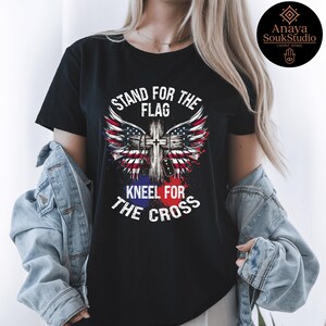 K&ouml;nnte beinhalten: Schwarzes T-Shirt mit dem Text "STAND FOR THE FLAG KNEEL FOR THE CROSS". Das Design zeigt ein Kreuz mit Fl&uuml;geln im amerikanischen Flaggenstil. Das T-Shirt hat einen klassischen Rundhalsausschnitt.