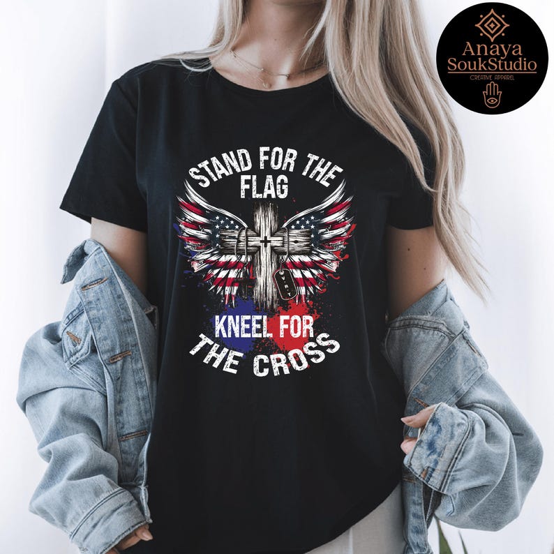 K&ouml;nnte beinhalten: Schwarzes T-Shirt mit dem Text "STAND FOR THE FLAG KNEEL FOR THE CROSS". Das Design zeigt ein Kreuz mit Fl&uuml;geln im amerikanischen Flaggenstil. Das T-Shirt hat einen klassischen Rundhalsausschnitt.