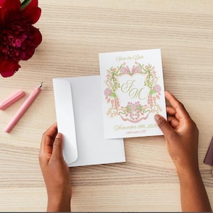 May include: A white envelope with a floral save the date card. The card has a pink and green floral design with the initials "J & M" in gold script. The text "Save the Date" and "November 11th, 2023" is printed in gold script.