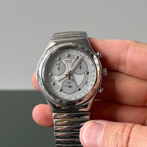 May include: A silver-toned Swatch watch with a white face and three sub-dials. The watch has a stainless steel band and a round bezel with black markings. The watch is held by a hand.