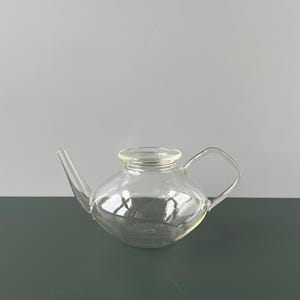 May include: A clear glass teapot with a rounded body, a spout, and a square handle. The teapot is designed for brewing tea and has a simple, elegant aesthetic. The glass allows the tea's colour to be visible.