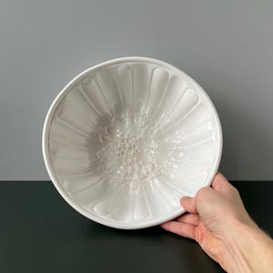 Op de afbeelding: Witte keramische vorm met een geribbeld ontwerp en een reliëfbloemmotief in het midden.