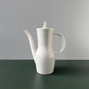 Könnte beinhalten: Eine weiße Kaffeekanne aus Keramik mit schlichtem, elegantem Design. Die Kanne hat einen runden Korpus und einen Ausguss auf einer Seite. Der Henkel ist an der Seite der Kanne befestigt.