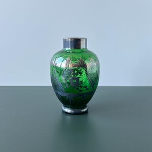Puede incluir: Un pequeño jarrón de vidrio verde con borde y base plateados. El jarrón presenta un diseño floral en un tono más oscuro de verde. El jarrón es un objeto decorativo.