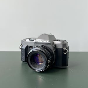 Può includere: Una fotocamera Pentax vintage, nera e argento. L'obiettivo è nero e presenta la scritta "SMC PENTAX-A". La fotocamera è posizionata su una superficie verde.