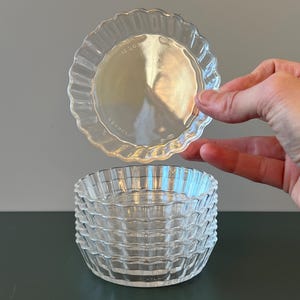 Puede incluir: Un conjunto de ramequines de vidrio transparente, acanalados, con borde festoneado. Un ramequín se sostiene por encima de la pila. Los ramequines son redondos y tienen un diseño acanalado. La imagen está tomada en interiores.