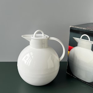 May include: A white, spherical, insulated carafe with a handle and a lid with a small handle. The carafe has a spout for pouring. A box with a similar image is partially visible in the background.