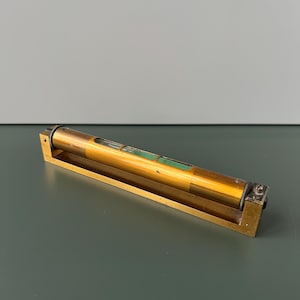 Puede incluir: Un nivel de burbuja de latón con tres viales de burbuja verdes. El nivel mide aproximadamente 15 cm de largo y tiene una base rectangular.