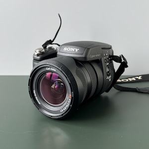 May include: Black Sony Cyber-shot digital camera with a Carl Zeiss lens. The camera has a retractable lens, a built-in flash, and a black strap. The lens has the text "Vario-Sonnar" and "2.8-4.8/4.3-71.5 T*" printed on it.
