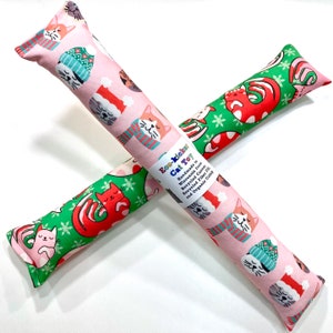 May include: Two cat toys with festive patterns. One is pink with cats wearing hats and scarves, the other is green with red cats and snowflakes. The toys are labeled "Eco-kickers Cat Toy" and are handmade in Wisconsin.