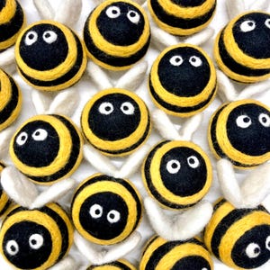 May include: A collection of handmade felt bee ornaments. Each bee features a black body with yellow stripes and white eyes. The bees have white felt wings, creating a whimsical and playful design.